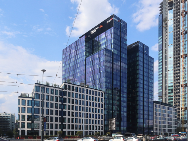 Großes Gebäude mit vielen Fenstern, die Deutsche Bank Hauptstadtsitz in Frankfurt, Deutschland, umgeben von städtischen Elementen einschließlich Straßenlaternen, Fahrzeugen, Bäumen und einem bewölkten Himmel.
