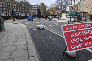 Eine Straße mit Fahrzeugen, ein Gehweg auf der linken Seite, Verkehrskegel, eine Ampel, ein Schild mit der Aufschrift "3-way control wait here until green light stops" und Menschen auf dem Gehweg, mit Bäumen, Gebäuden und einem bewölktem Himmel im Hintergrund.