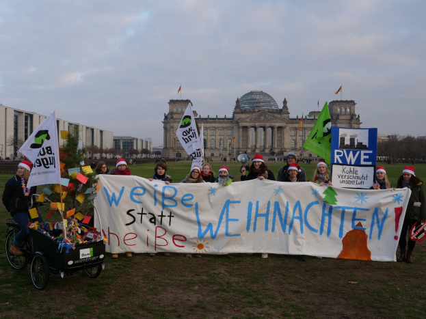 Gruppe von Menschen in Mützen mit einem Banner vor dem Reichstag, mit einer Person in einem Kinderwagen, grasbewachsenem Boden, Bäumen, Gebäuden und Fahnenmasten im Hintergrund bei bewölktem Himmel.