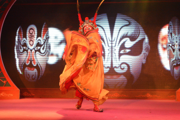 Ein Darsteller in Kostüm und Maske führt auf der Bühne eine dramatische Pose aus, während auf dem Hintergrund projizierte Bilder zu sehen sind.