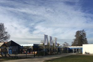 Ein großes Gebäude mit einem grasbewachsenen Bereich davor, umgeben von einem Metallzaun, einem Pfad, Bäumen, Pfosten, Bannern, einem geparkten Fahrrad, benachbarten Häusern und einem bewölkten Himmel, das als neuer Spielbereich für Kinder an einer Schule dient.