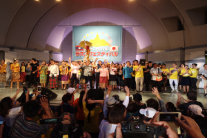 Eine Gruppe von Menschen auf einer Bühne, einige halten Kameras und Handys, andere Musikinstrumente, mit einem Banner und Lichtern im Hintergrund, was eine Festivaleröffnung vermuten lässt.