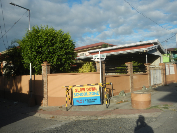 Schulzonen-Schild mit der Aufschrift "Slow Down School Zone" vor einem Gebäude mit Bäumen, Polen, Lampen, Drähten und einer Wand unter einem bewölkten Himmel.