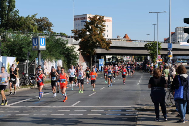 Gruppe von Menschen, die bei einem Marathon auf einer baumbestandenen Straße mit Laternen, Schildern, Fahrrädern, einem Zaun, Gras, einer Brücke, Gebäuden und einem klaren blauen Himmel laufen.