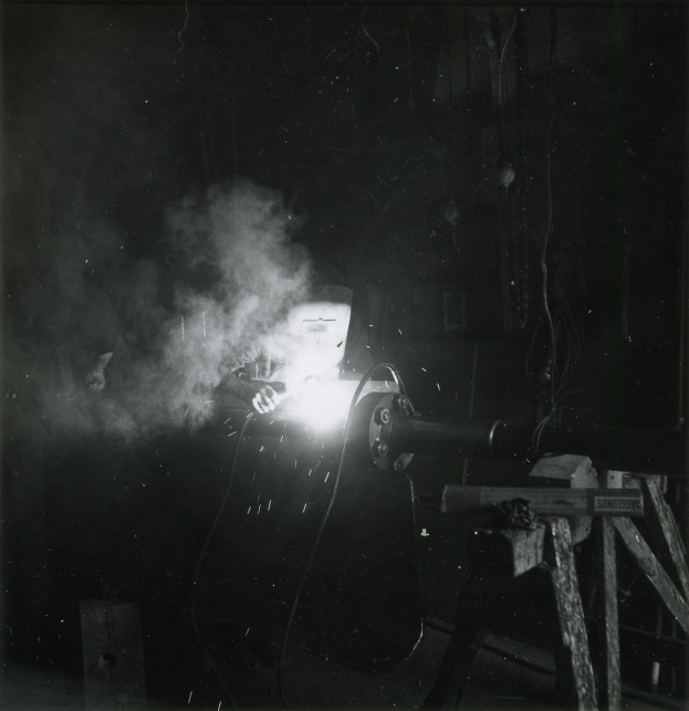 Schwarzes und weißes Foto eines Schweißers bei der Arbeit an einem Metallstück, mit Rauch aus der Schweißmaschine und verschiedenen Gegenständen im Hintergrund.