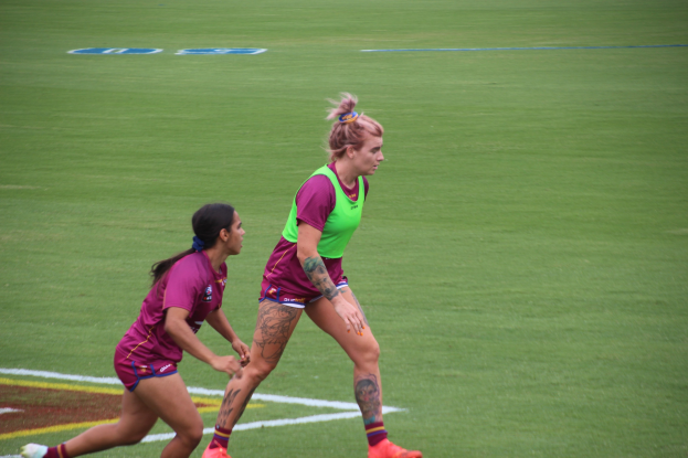 Zwei Frauen spielen Fußball auf einem Feld, eine trägt ein grünes T-Shirt und Shorts, die andere ein pinkes Kleid, beide tragen Sportkleidung und haben sichtbare Tattoos an Händen und Füßen.