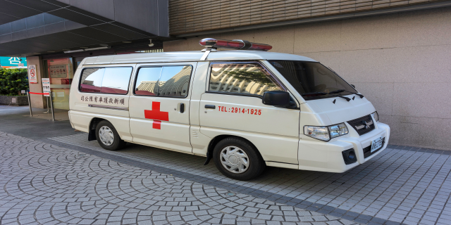 Ein Rettungswagen steht vor einem Gebäude mit einer Tafel, umgeben von Bäumen und Pflanzen.