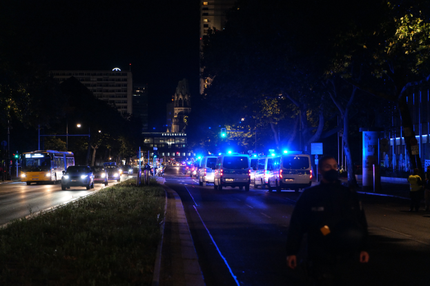 Eine nächtliche Stadtstraße mit Polizeiwagen, Bussen, Fußgängern auf dem Gehweg, Gras links, Bäumen, Gebäuden und beleuchteten Laternen.