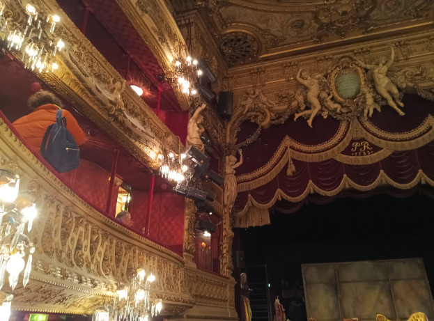 Theaterinnenraum mit Kronleuchtern, Wandskulpturen und Menschen auf der Bühne, mit verstreuten Gegenständen, die eine lebendige Atmosphäre schaffen.