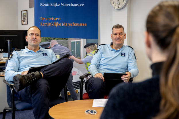 Zwei Polizeibeamte sitzen an einem Tisch mit Papieren, sprechen mit einer Frau; eine wandmontierte Flagge, Uhr und Fotorahmen sind im Hintergrund zu sehen, sowie ein weiterer Tisch mit Monitor, Tastatur und Maus links daneben.