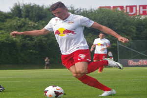 Professioneller Fußballspieler, der einen Ball auf einem Rasenfeld mit "RB Leipzig"-Schriftzug im Hintergrund unter einem klaren blauen Himmel tritt.