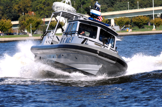 Ein Polizeiboot fährt über einen Gewässer mit einer Person darin, mit einer Brücke, Bäumen, Polen, Fahrzeugen, Gras und einem klaren blauen Himmel im Hintergrund.