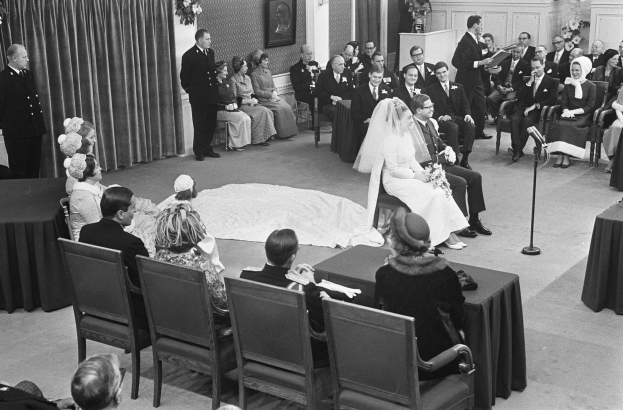 Schwarzes und weißes Foto einer Hochzeitszeremonie mit Menschen, die sitzen und stehen, einem Mann mit Mikrofon, einer Frau mit Blumenstrauß, einem Podium mit Mikrofon, Blumenschmuck, einem Vorhang, einem Rahmenfoto an der Wand und einer Tür im Hintergrund.