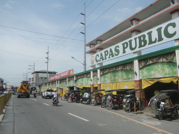 Eine belebte Stadtstraße mit Fahrzeugen, einem Geländer auf der linken Seite, Gebäuden, Polen, Laternen, Schildern und einigen Menschen unter einem bewölkten Himmel, mit einem Gebäude mit der Aufschrift 'Capas Public'.