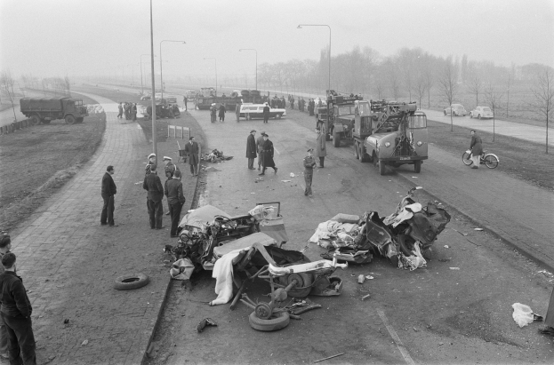 Schwarz-weiß-Bild eines Autounfalls am Straßenrand mit mehreren Fahrzeugen, einer Gruppe von Menschen in der Nähe und Laternen, Bäumen und Himmel im Hintergrund.