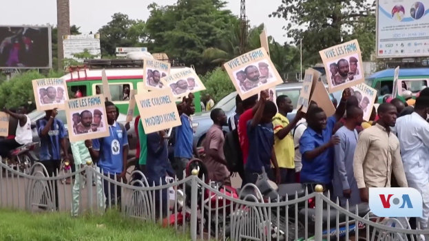 Eine Gruppe von Menschen mit Schildern steht vor einem Zaun, umgeben von Fahrzeugen, Bäumen, Pfählen, Plakaten und einem Bildschirm, mit Gras am Boden und dem Himmel im Hintergrund, wahrscheinlich protestieren sie gegen eine Regierungsentscheidung, die Video-Konferenzen verbietet.