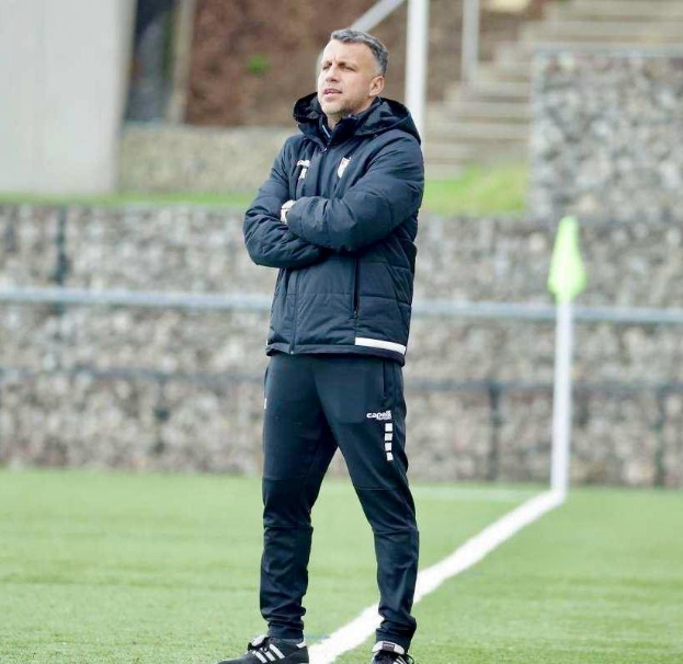 Ein Fußballtrainer auf einem Feld mit verschränkten Armen, der eine schwarze Jacke und Hose trägt, während eines Trainings.