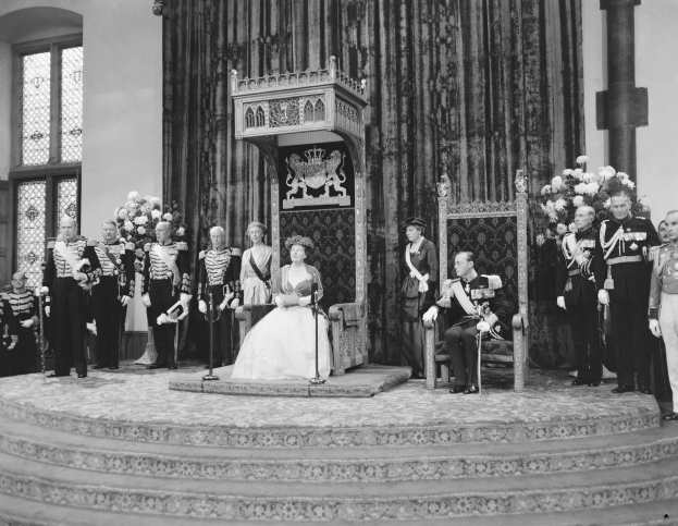 Schwarz-weißes Foto von Queen Elizabeth II., die auf einem Stuhl sitzt und ein weißes Hochzeitskleid trägt und einen Blumenstrauß hält, neben Prinz Philip, Duke of Edinburgh, der rechts von ihr sitzt, und Begleitpersonen, die links von ihr stehen.