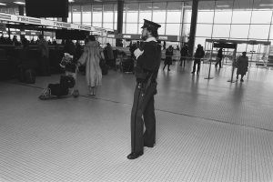 Ein Polizeibeamter in Uniform steht in einem Flughafen-Terminal und hält eine Waffe, umgeben von Menschen und Gepäckwagen, mit Säulen, Texttafeln, Deckenleuchten und Glaswänden im Hintergrund.