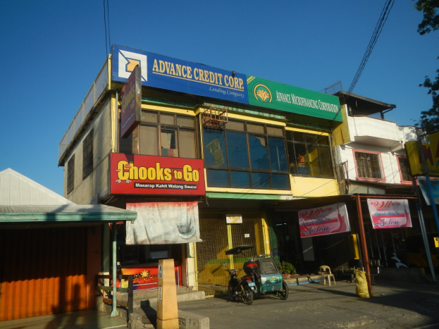 Ein Gebäude mit einer Tafel, die "Advance Credit Corp" trägt, auf einer Straße mit Fahrzeugen, Werbetafeln, Strommasten, Kabeln, Bäumen und einem klaren blauen Himmel.