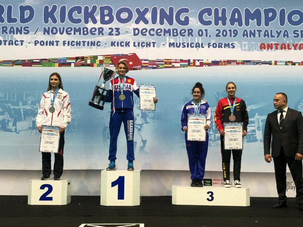 Fünf Personen mit Trophäen und Urkunden auf einem Podium vor einem "World Kickboxing Championship Antalya 2019"-Plakat im Hintergrund.