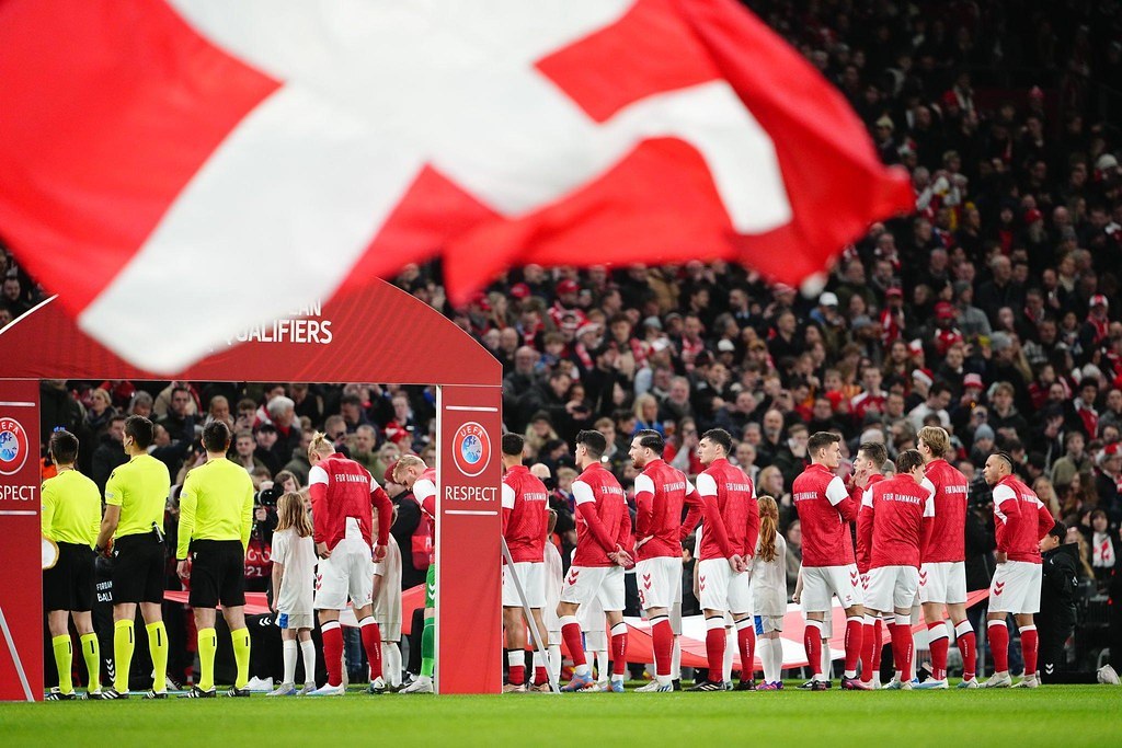 Gruppe von Menschen auf einem Fußballfeld mit einer roten und weißen Flagge im Vordergrund, einem Bogen mit "Bayern Munich vs Bayern Munich Betting Tips & Preview"-Text im Hintergrund und einer großen Menschenmenge im Stadion.