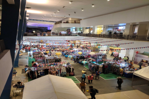 Eine feierliche Menge schlendert durch einen belebten Indoor-Weihnachtsmarkt im Mall of Asia mit Ständen, aufgehängten Kleidungsstücken, dekorativen Fahnen, Ballons, sitzenden und stehenden Menschen, Geländern, Säulen und Deckenlampen.