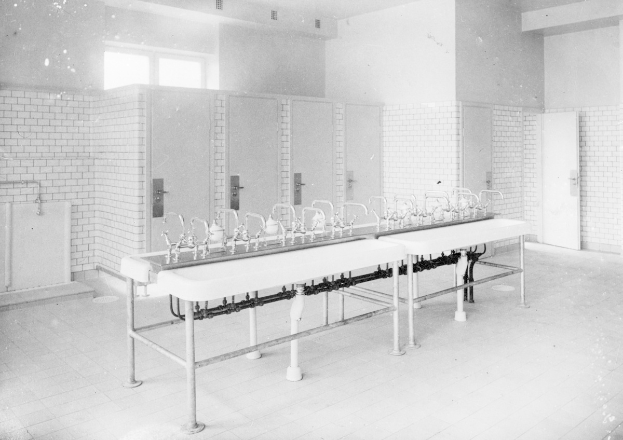 Schwarz-weiß-Foto einer öffentlichen Toilette mit einer Reihe von Waschbecken, Wasserhähnen, Wänden, Türen, Fenstern und einem gefliesten Boden, auf dem Gegenstände verstreut sind.