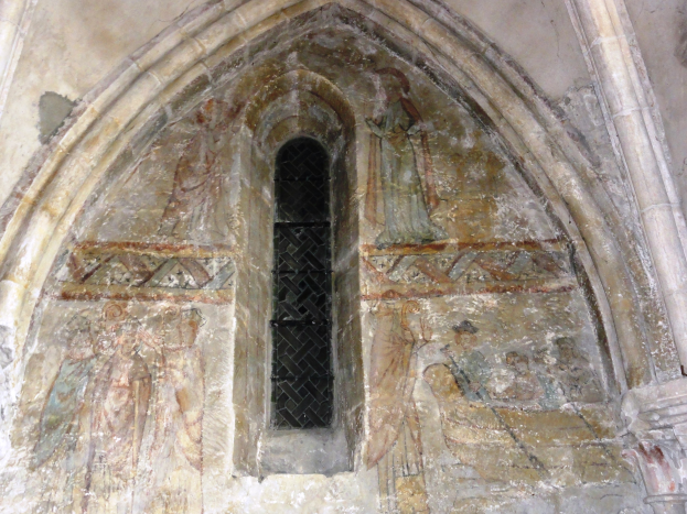 Ein alter Kircheninnenraum mit einem steinernen, bögenförmigen Fenster und einer farbenfrohen Fresken-Schmuckwand mit detaillierten Entwürfen und verschiedenen Szenen und Figuren.