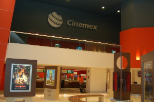 Eine Gruppe von Menschen steht in einem Cinemex-Kino in einem Einkaufszentrum, mit einer Person, die sitzt, Tischen, Fotorahmen an der Wand, einem Schild, Deckenleuchten und Geräten auf den Tischen.