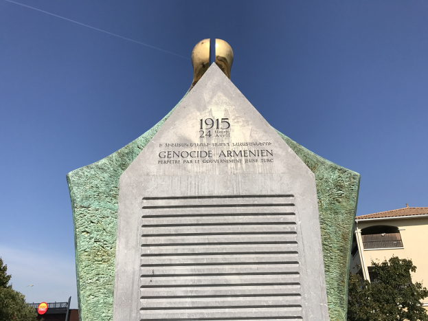 Ein steinernes Denkmal mit einer Uhr darauf, umgeben von Bäumen und Gebäuden, mit Inschrift zum Gedenken an den 24. Jahrestag des Völkermords in Armenien.