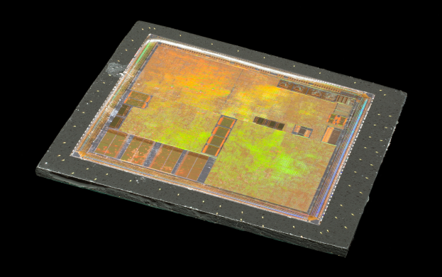 Nahaufnahme eines rechteckigen Halbleiterchips mit glänzender Oberfläche, kleinen zentralen Löchern und einem schmalen Metallrahmen vor schwarzem Hintergrund.