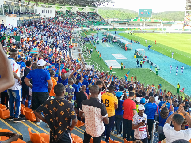 Eine große Menschenmenge versammelt sich vor einem eingezäunten Fußballfeld, einige sitzen und andere stehen, unter einem klaren blauen Himmel mit Hügeln im Hintergrund, und warten auf das Spiel der 2019 African Cup of Nations.