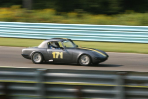 Ein Auto mit einem Helm tragenden Fahrer bewegt sich auf einer Straße, die von Barrieren gesäumt ist, mit Bäumen im Hintergrund.