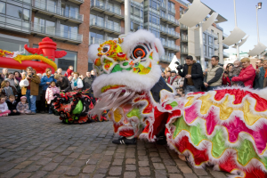 Ein lebendiges chinesisches Neujahrsfest in Amsterdam mit einer Löwen-Tanzvorstellung und einer Menge Schaulustiger, einige mit Kameras, vor einem Hintergrund aus Gebäuden, Laternenmasten und einem klaren blauen Himmel.