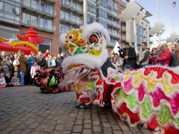 Ein lebendiges chinesisches Neujahrsfest in Amsterdam mit einer Löwen-Tanzvorstellung und einer Menge Schaulustiger, einige mit Kameras, vor einem Hintergrund aus Gebäuden, Laternenmasten und einem klaren blauen Himmel.