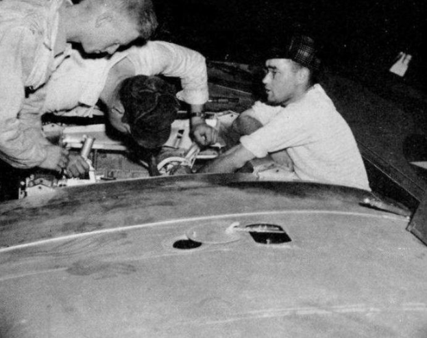 Drei Männer mit Mützen arbeiten an einem schwarz-weißen Auto in einer Garage, umgeben von Werkzeugen und Materialien.