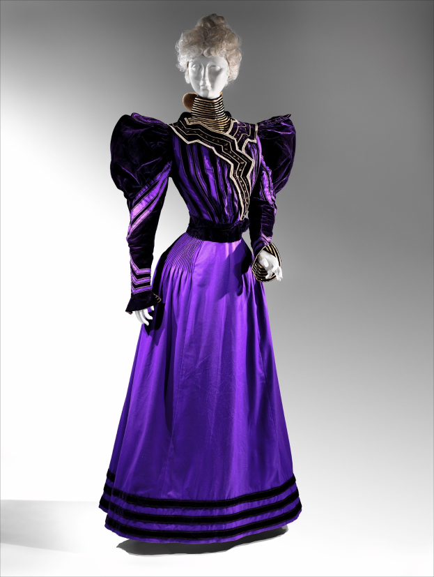 Eine Schaufensterpuppe in einem lila Abendkleid mit schwarzem und goldenem Muster, das eine eng anliegende Taille, einen langen Rock bis zum Boden mit goldener Stickerei und Perlen und eine Spitzenverzierung an der Taille hat, kombiniert mit einem eleganten Hochsteckdutt und zarten Ohrringen.