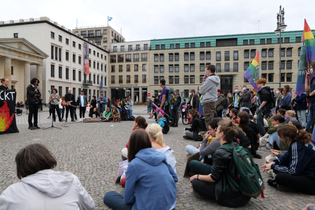 Eine Gruppe von Menschen, die auf dem Boden vor einer Menge mit Fahnen und Transparenten sitzen, mit einer Person, die in ein Mikrofon spricht, einer Statue und Gebäuden im Hintergrund während einer anti-schwulen Demonstration in Berlin.