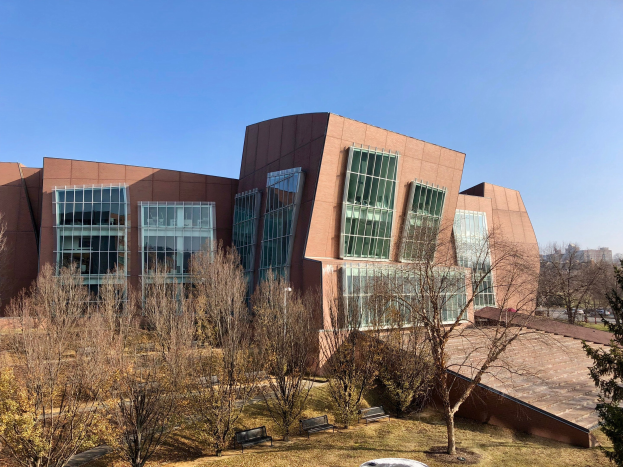 Großes Gebäude mit vielen Fenstern, umgeben von Bäumen, Bänken, Gras und einem Laternenpfahl, mit anderen Gebäuden und einem klaren blauen Himmel im Hintergrund, als der neue Campus der University of Illinois at Chicago identifiziert.