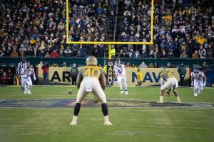 Fußballspieler in Sportkleidung und Helmen konkurrieren auf einem Feld mit Zuschauern in den Rängen, Polen und Tafeln mit Text.