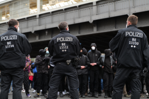 Eine Gruppe von Polizisten in Uniform steht vor einer Menge von Menschen in schwarzen Uniformen und Masken, mit einer ├╝berquerung und einem Geb├Ąude im Hintergrund, was auf eine Stadt als Schauplatz hindeutet.