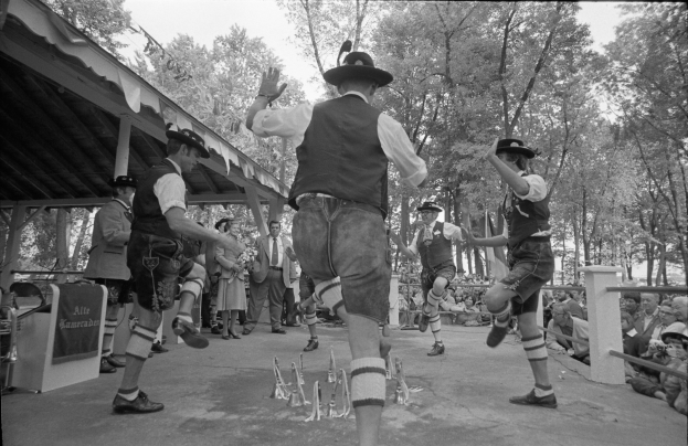Eine Gruppe von Männern in traditioneller bayrischer Tracht tanzt auf einer Bühne vor einem gemischten Publikum aus sitzenden und stehenden Menschen, mit Bäumen und einem Schuppen im Hintergrund, dargestellt in Schwarz-Weiß.
