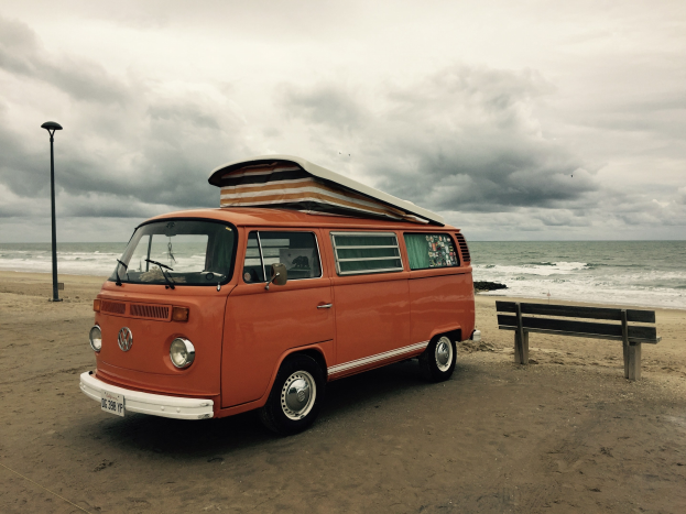 Orange Volkswagen-Bus auf einem Strand in der N├Ąhe des Meeres geparkt, mit einer Bank und einem Laternenpfahl in der N├Ąhe, unter einem bew├Âlktem Himmel.