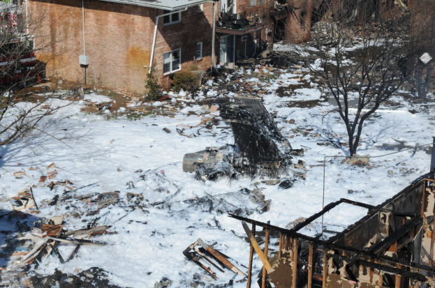 Verbrannte Überreste eines durch Feuer zerstörten Hauses in einer verschneiten Wohngegend, mit Bäumen und Gebäuden im Hintergrund.