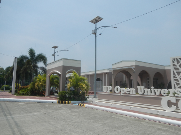 Der Eingang zu einem offenen Universitätscampus mit einer Straße, die zum Gebäude führt, umgeben von Bäumen und Pflanzen, einer Wand mit Text auf der rechten Seite und dem Himmel im Hintergrund.
