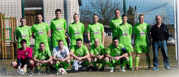 Eine Gruppe von Männern in grünen T-Shirts steht auf einem Fußballfeld, einige in Hockstellung, mit einem Ball vor ihnen und einem Torpfosten rechts daneben, vor einer Kulisse aus Bäumen, Gebäuden und Fahrzeugen.