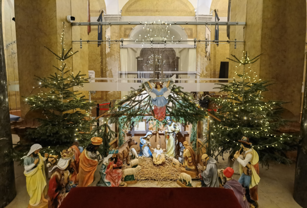 Eine Krippe in einer Kirche mit einem roten Tuch auf einem zentralen Tisch, flankiert von zwei beleuchteten Weihnachtsbäumen und einer Kronleuchter, die über einer Brüstung in den Hintergrund hängt.