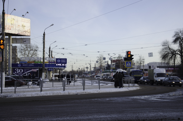 Eine belebte Stadtstraße mit starkem Verkehr an einem verschneiten Tag, mit Autos, Lastwagen, Straßenlaternen, Geländern, Polen, Brettern, Drähten, Bäumen, Gebäuden und Fußgängern unter einem bewölkten Himmel.
