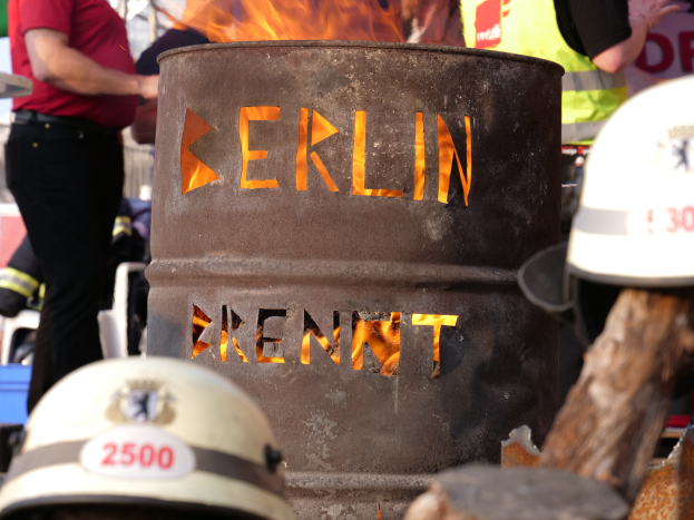 Eine Gruppe von Menschen um einen Feuerhydranten mit der Aufschrift "Berlin" herumstehend mit einem brennenden Fass im Vordergrund, Helme unten und ein hölzernes Objekt auf der rechten Seite.